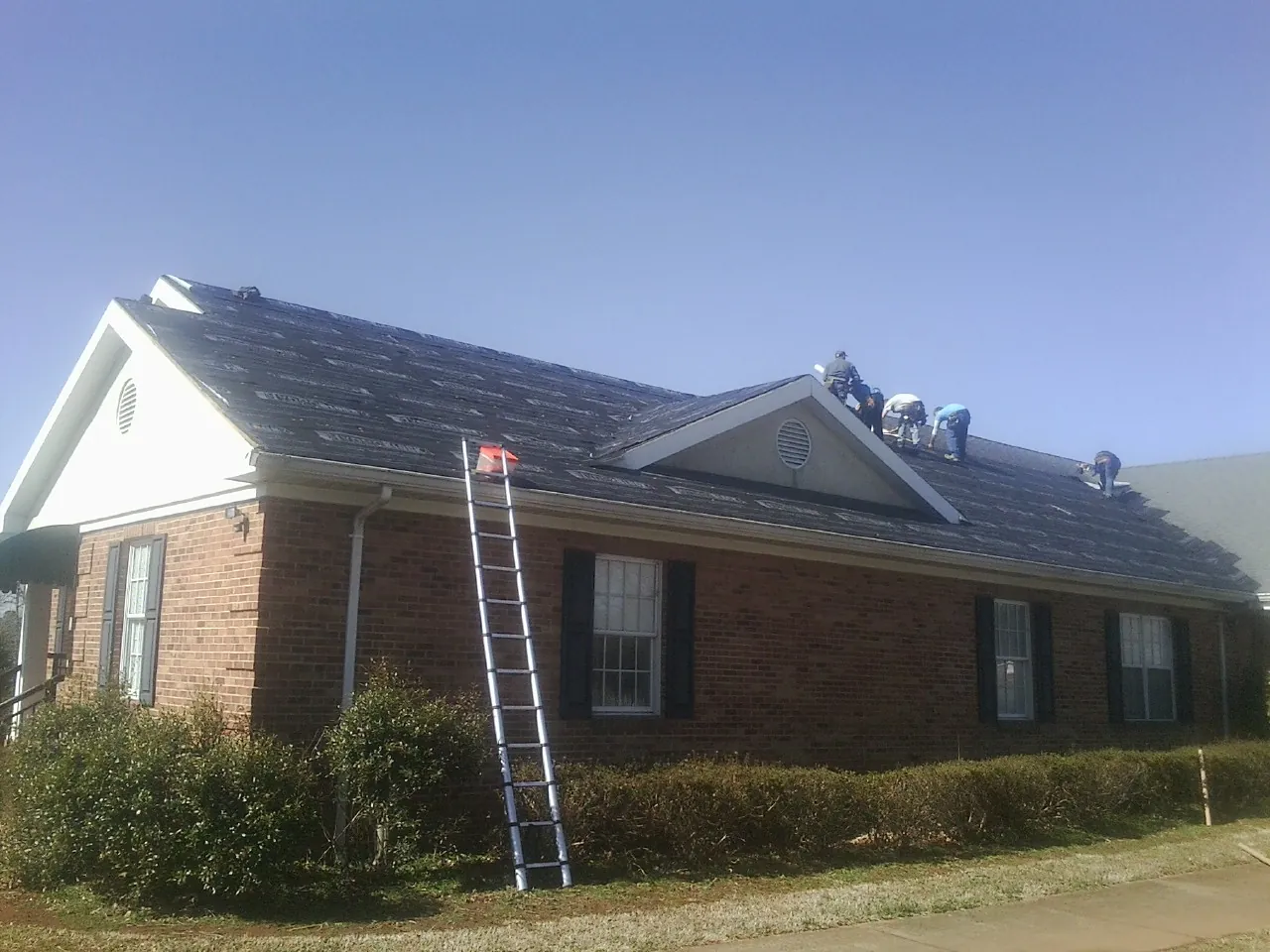 Crew tearing shingles off a brick ranch roof with a ladder set — Metal Roof Repair work by our crew in Chattahoochee