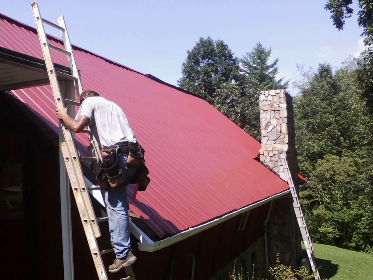 Roofer on a ladder beside a red metal roof and stone chimney — Soffit & Fascia Repair by Roofing Brothers in Chattahoochee